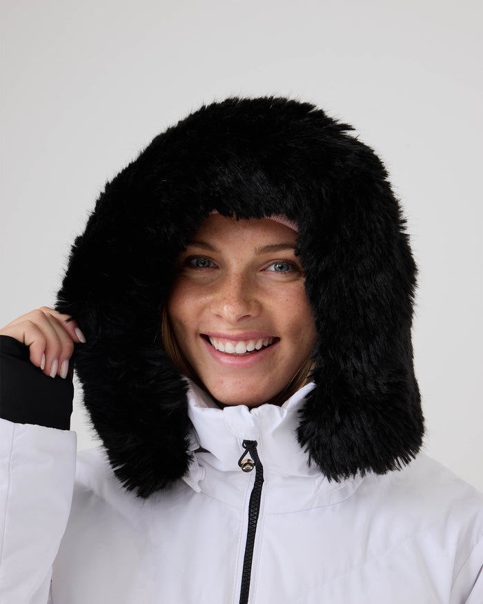 A person wearing the Roxy Snowdrift Jacket—a white women’s ski jacket featuring a large, fluffy black faux-down hood—smiles at the camera while holding the hood with one hand. The background is plain and light-colored.