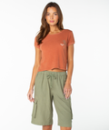 A woman stands facing forward against a plain white background, wearing the Roxy Spanish Summer Cropped Baby Tee in Chutney Spanish Summer—a rust-colored, breezy-fit top—paired with olive green cargo shorts featuring a drawstring and pockets.