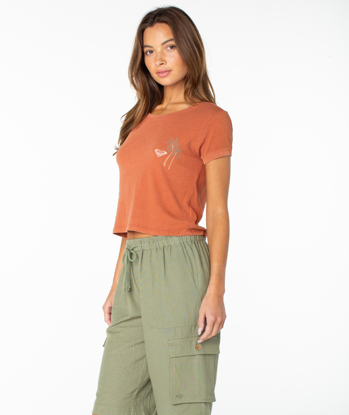 A woman wearing the Roxy Spanish Summer Cropped Baby Tee—an orange short-sleeve graphic tee with a small palm tree and shell design—paired with green cargo pants, stands against a plain white background.