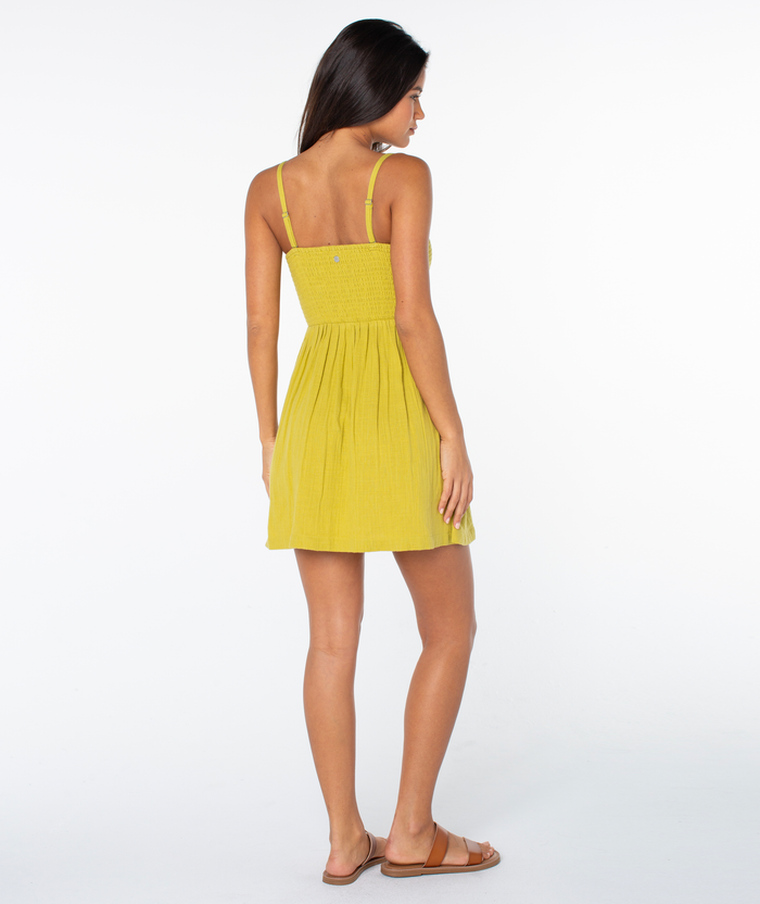 A woman faces away, wearing the Roxy Spring Fling Mini Dress in light yellow with thin straps and brown sandals, set against a plain white background.