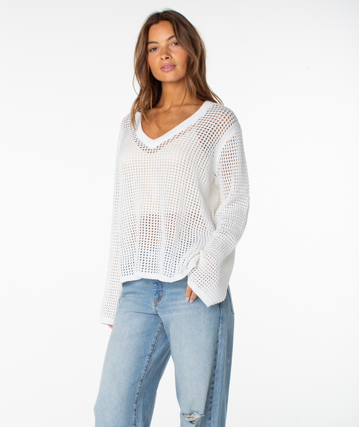 A woman stands against a white background wearing the Roxy Summer Nights V Neck Tunic over a white top and light blue jeans, hand in her pocket and looking relaxed—an ideal summer staple.