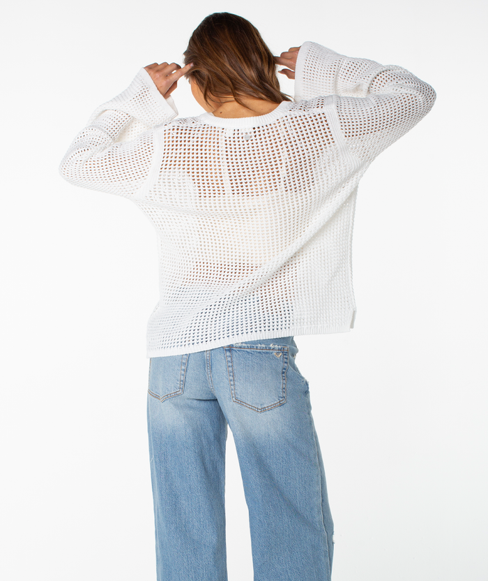 A person with brown hair wears the Roxy Summer Nights V Neck Tunic—a loose, white cotton sweater—paired with light blue jeans, adjusting the collar. The plain white background highlights this effortless summer staple.
