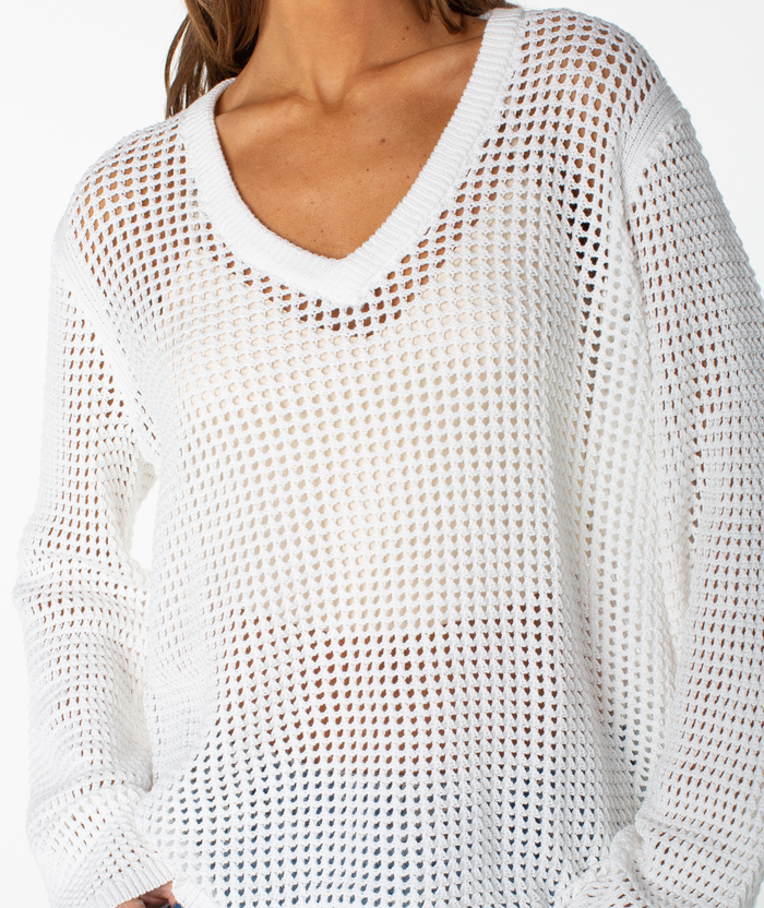 A person wears the Roxy Summer Nights V Neck Tunic, a white, long-sleeve cotton sweater with a mesh-like, see-through pattern. This loose-knit piece is an ideal summer staple. The person's head is not visible.