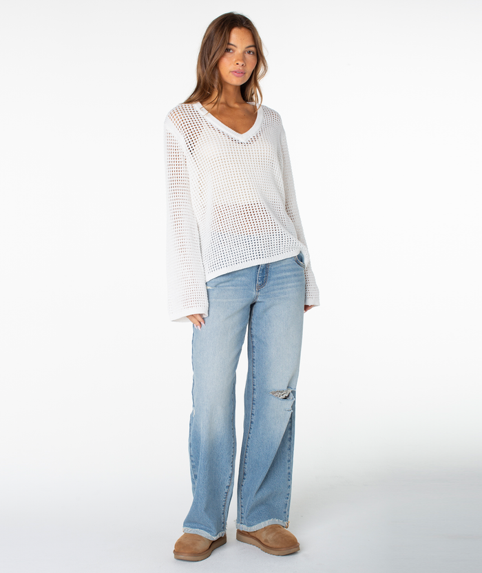A woman with long brown hair poses casually against a white background wearing the Roxy Summer Nights V Neck Tunic sweater over a white top, paired with light blue distressed jeans and tan slippers.