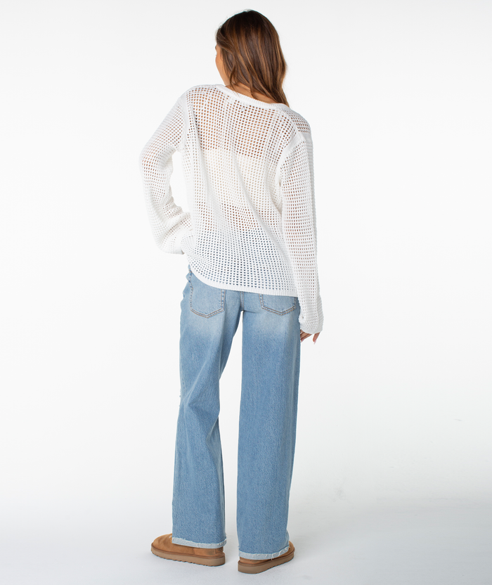 A woman faces away, wearing the Roxy Summer Nights V Neck Tunic sweater in white, paired with light blue wide-leg jeans and tan slippers, set against a plain white background.