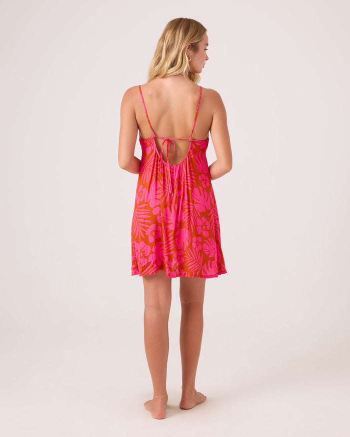 A woman stands barefoot facing away, wearing the Roxy Sun Chasers Plunge Dress Cover Up—a vibrant pink tropical-print piece with thin straps, open back tied in a bow, and textured crinkle rayon fabric against a light background.