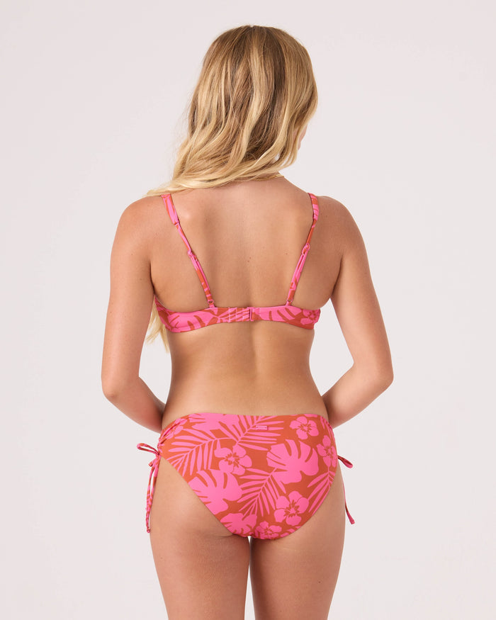A woman with long blonde hair is seen from the back, wearing the Roxy Sun Chasers Scoop Bralette Bikini Top with adjustable straps and side-tie bottoms in pink tropical floral, standing against a plain light background.