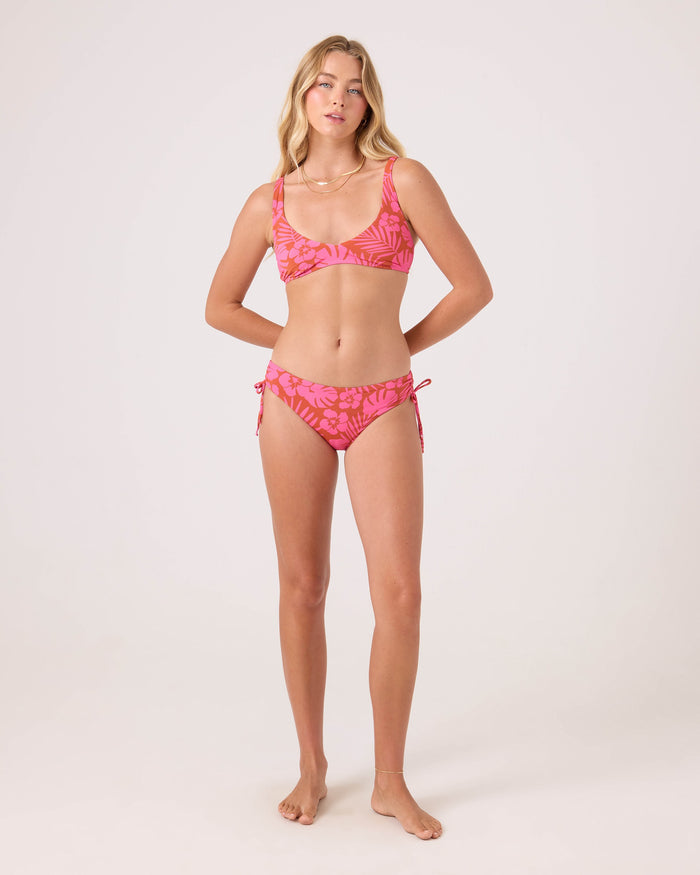 A woman with long, wavy blonde hair stands barefoot against a plain background, wearing the Roxy Sun Chasers Scoop Bralette Bikini Top in pink with a leafy pattern and adjustable straps. Her arms are relaxed at her sides.