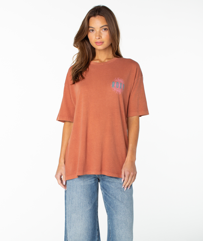 A woman stands against a plain white background, wearing the Roxy Sun Club Oversized Tee—an oversized, rust-colored shirt with relaxed drop shoulders and a small chest graphic—paired with blue jeans.