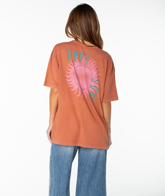 A person with long brown hair, wearing the Roxy Sun Club Oversized Tee—a relaxed, drop-shoulder, rust-colored t-shirt featuring a colorful sun and text design on the back—and blue jeans stands facing away against a white background.