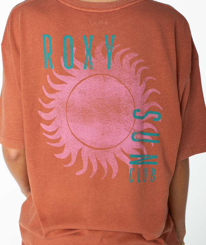 Wearing the Roxy Sun Club Oversized Tee in a rust hue, featuring a large pink sun graphic and turquoise “ROXY SUN CLUB” text on the back, this lightweight top offers a relaxed drop shoulder for an effortlessly cool look.