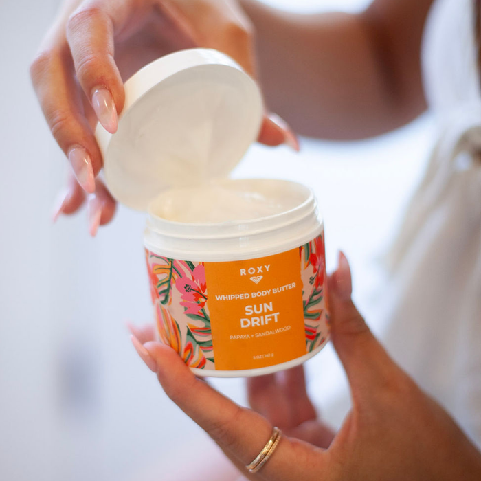 A person with manicured nails holds an open jar of Roxy Sun Drift Body Butter with a vibrant floral label—ideal for daily hydration and soothing dry skin.