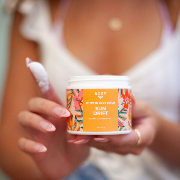 Someone holds a jar of Roxy Sun Drift Body Scrub with its vibrant floral label, while their other hand shows a scoop of the scrub. The blurred background emphasizes this Roxy self-care moment.