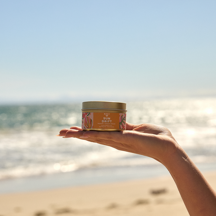 A hand holds the Roxy Sun Drift Palm Tin Candle against a sunny beach with blue sky and sparkling ocean—an ideal tropical-scented candle for relaxing by the seaside.