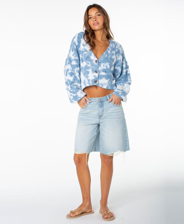 A woman stands against a white background, wearing the Roxy Sundaze Sweater—a blue dip-dye knitwear cropped sweater with a button front—paired with light denim knee-length shorts featuring a raw hem and beige flip-flops. Her wavy hair frames her face as she casually slips her hands into her pockets.