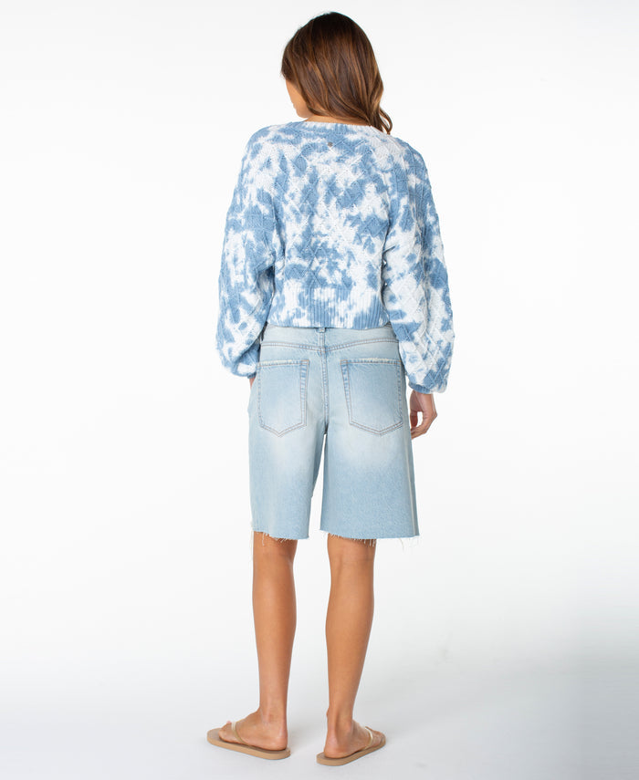 A woman stands facing away against a plain white background, wearing the Roxy Sundaze Sweater—a blue and white dip-dye knitwear piece—paired with light-wash denim Bermuda shorts and tan slide sandals.