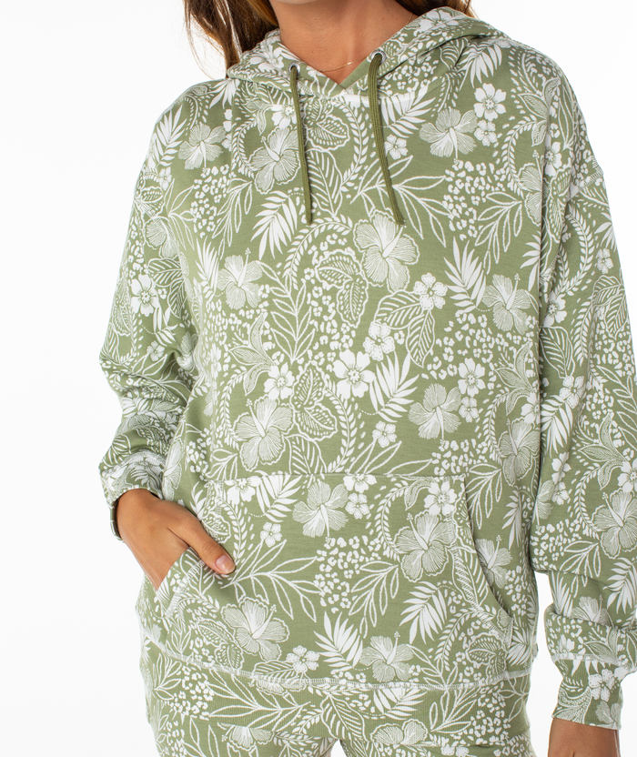 A person wearing the Roxy Surfing By Moonlight Printed Hoodie in light green with a white floral and leafy design stands with one hand in the front pocket. The image is cropped at the shoulders and mid-thigh.