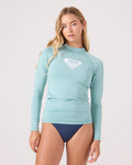 A woman with long blonde hair models the Roxy Whole Hearted Long Sleeve Rashguard in light blue, featuring a white chest logo and made from recycled fabric, paired with navy bikini bottoms against a plain light background.