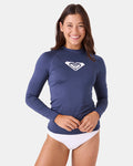 A woman with long brown hair smiles while wearing the Roxy Whole Hearted Ls Rashguard in navy blue, made from recycled polyester with a white logo on the chest, paired with white swim bottoms, against a plain white background.