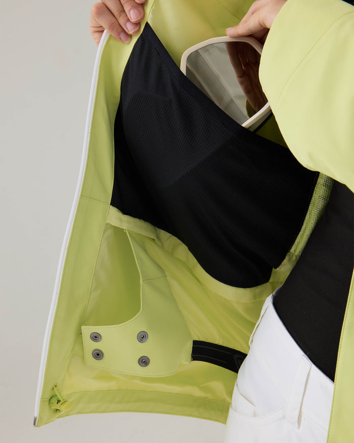 A woman opens the light green Roxy Zephyr Jacket, revealing an interior black mesh pocket holding a pair of white goggles. Designed for ski and snowboard adventures, this technical outerwear is paired with white pants and a black shirt.
