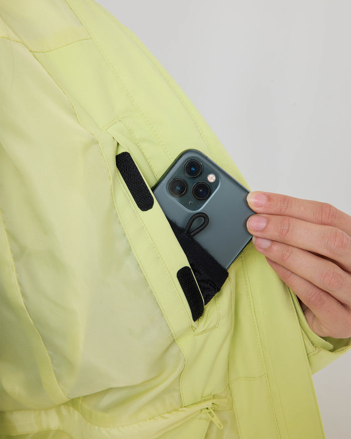 A hand places a smartphone with three rear cameras into the inner pocket of the light yellow Roxy Zephyr Jacket, showcasing the technical outerwear’s practical design that makes it a favorite for ski and snowboard adventures.