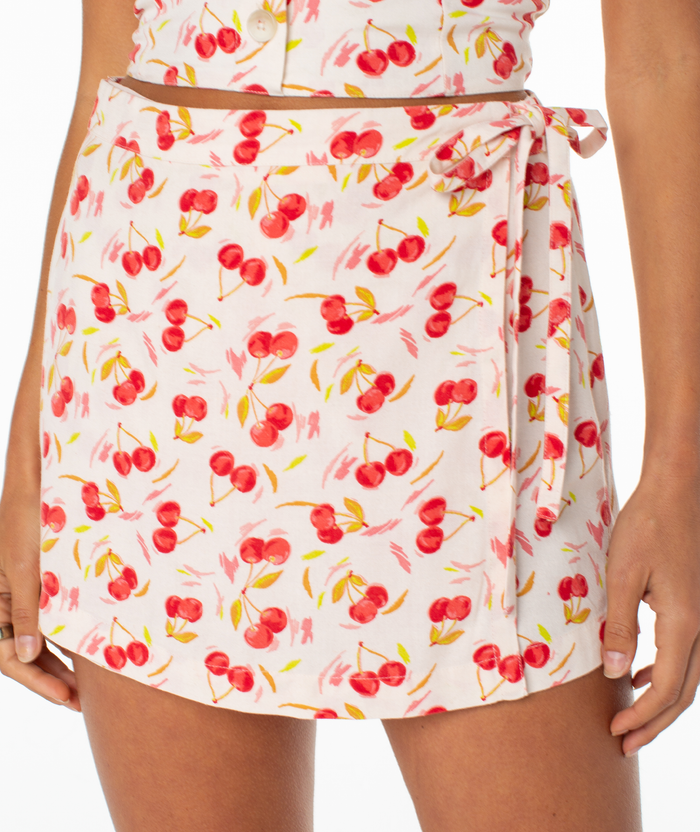 A person wears a white Roxy Zola Skort with a red cherry and floral print, featuring a side tie waist and bow. Only the lower torso and upper legs are shown. The matching top is also visible.